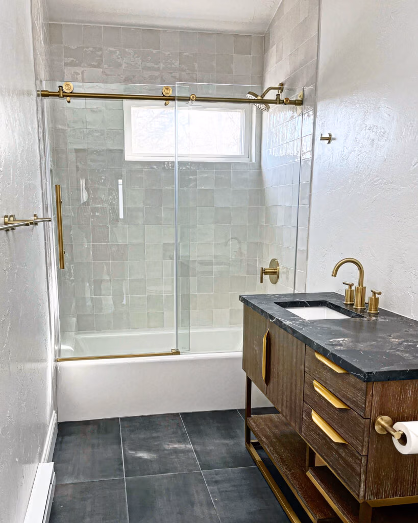 Modern bathroom design featuring a glass shower enclosure, gold fixtures, a dark stone countertop vanity, and gray tiled flooring, showcasing high-quality materials ideal for Summit County remodeling.