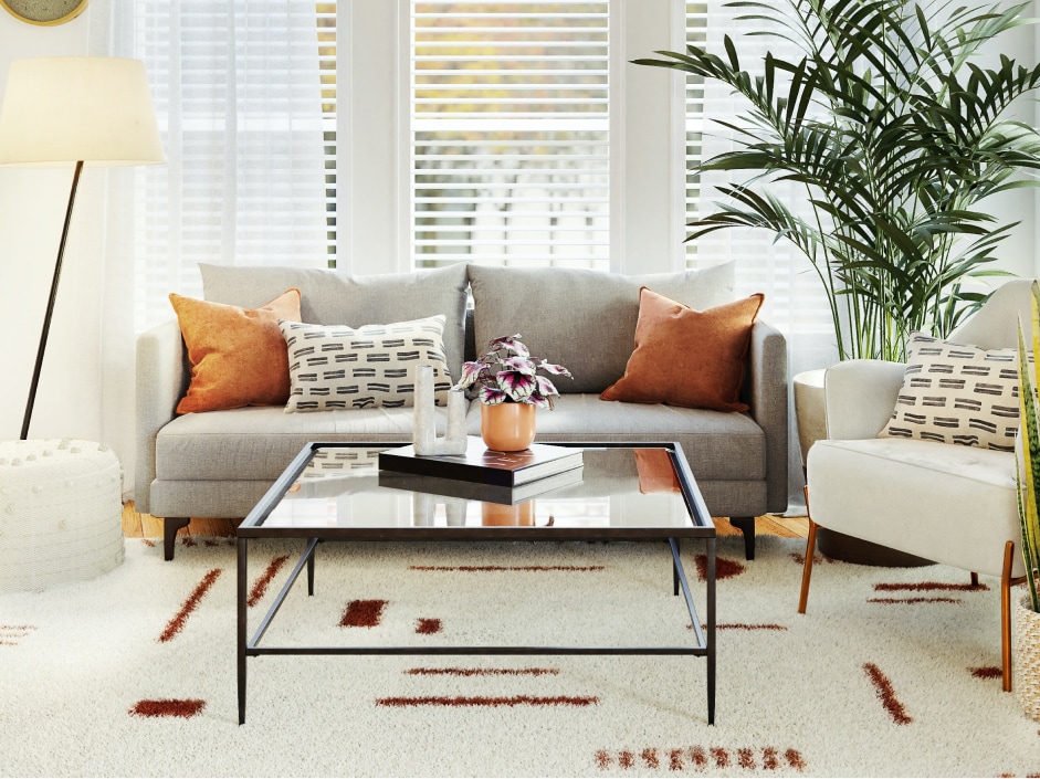 Cozy living room interior featuring a gray sofa with decorative pillows, a glass coffee table with a plant centerpiece, and a bright atmosphere enhanced by natural light from large windows.
