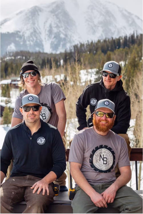 Four team members of Natural Builders posing outdoors in a mountainous landscape, wearing branded shirts and caps, showcasing a collaborative and energetic work culture.